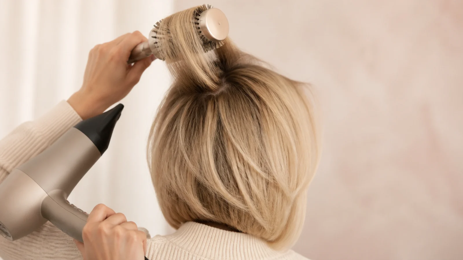 A round brush lifting a section of blonde hair at the root under a hairdryer